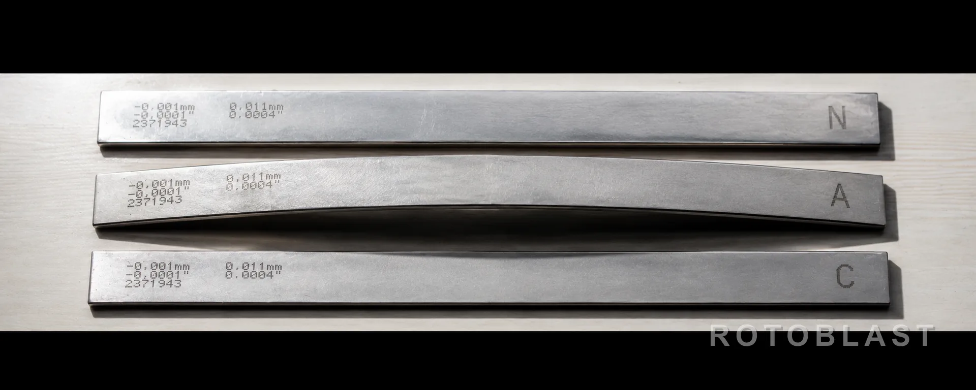 SUBJECT: three Almen strips (N, A, C) laid side by side on a clean surface, one visibly peened and arched. FRAMING: ultra-wide letterbox echoing the strip's own 4:1 proportion; strips running the full width. LIGHTING: hard raking light from one side so the arch of the peened strip throws a readable shadow. ANGLE: near-flat, slightly above, almost elevation. PURPOSE: establishes that this is precision stock, not sheet metal — and shows the deflection that the whole page is about.