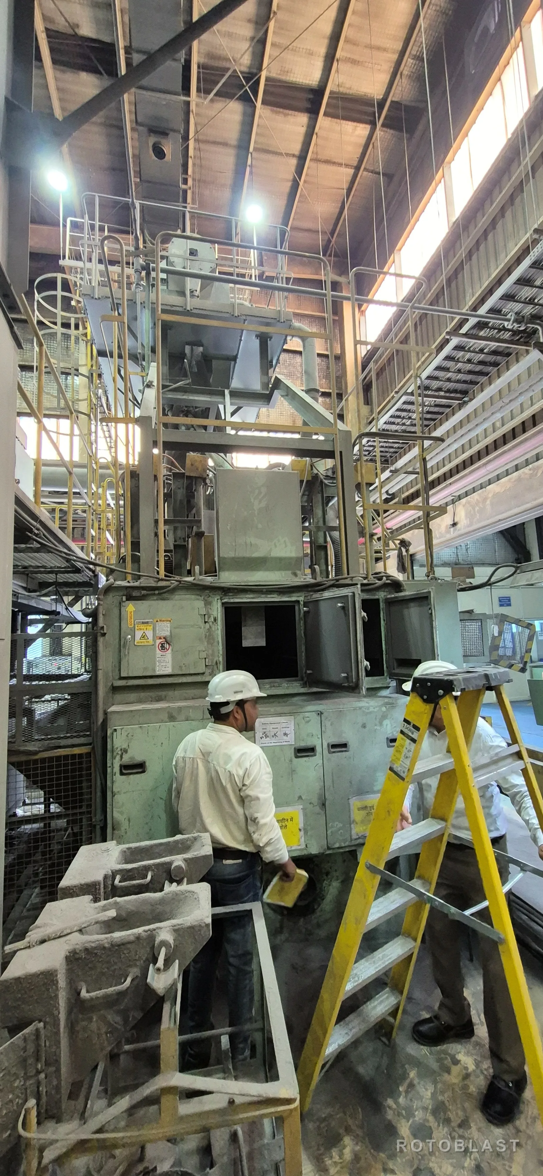 Engineers in hard hats working on an installed Rotoblast shot blasting machine at a customer's plant, blast cabinet doors open and castings staged on a trolley in the foreground.