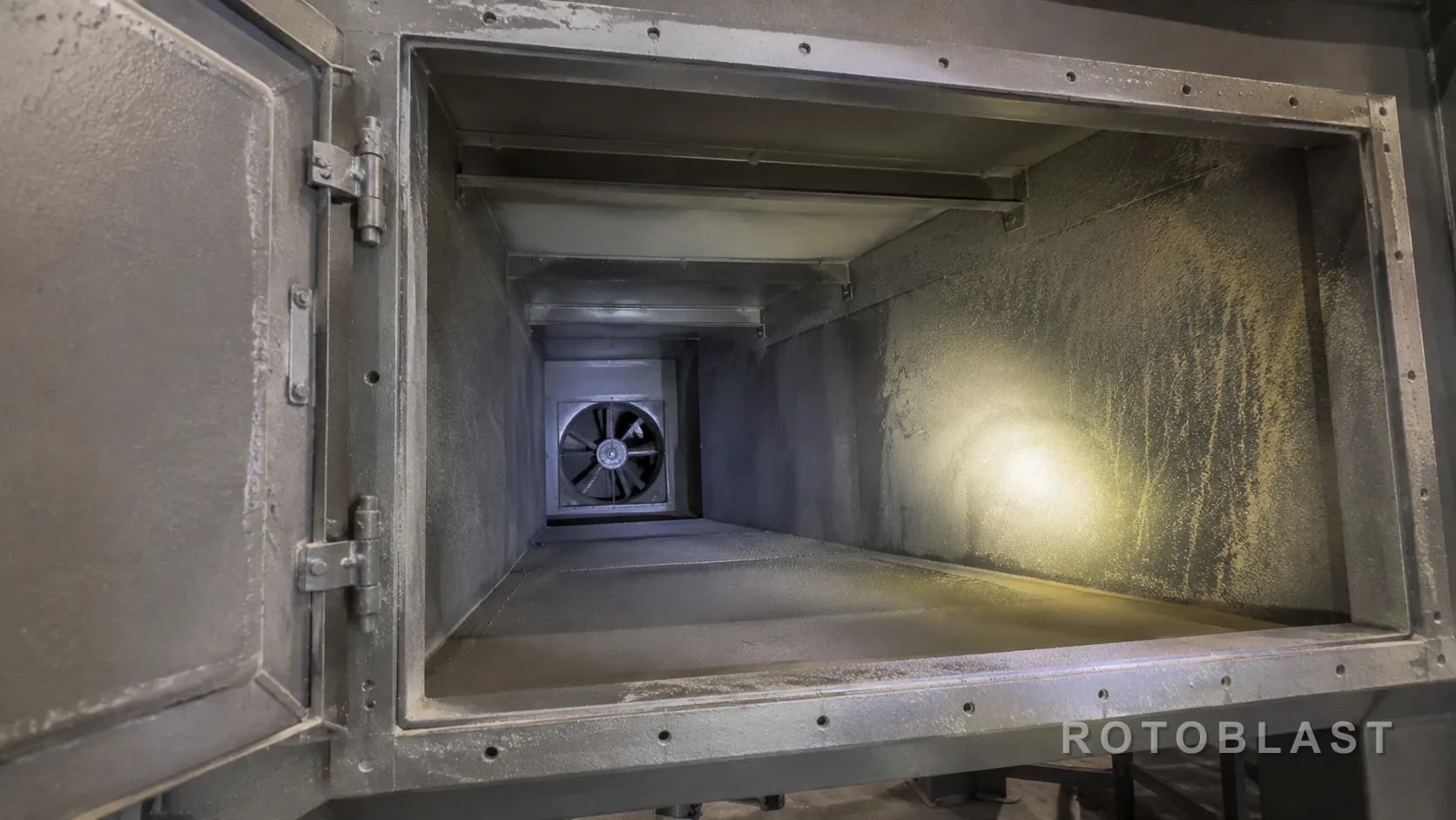 SUBJECT: the clean-air plenum of a dust collector with the access door open, showing dust deposited where there should be none. FRAMING: 16/9, looking into the plenum toward the fan inlet. LIGHTING: available light plus a torch beam picking out the dust film on a surface that should be clean. ANGLE: standing at the open door. ENGINEERING STORY: dust on the clean side is the proof of a filter fault. PURPOSE: the reader can go and check this on their own machine in five minutes.