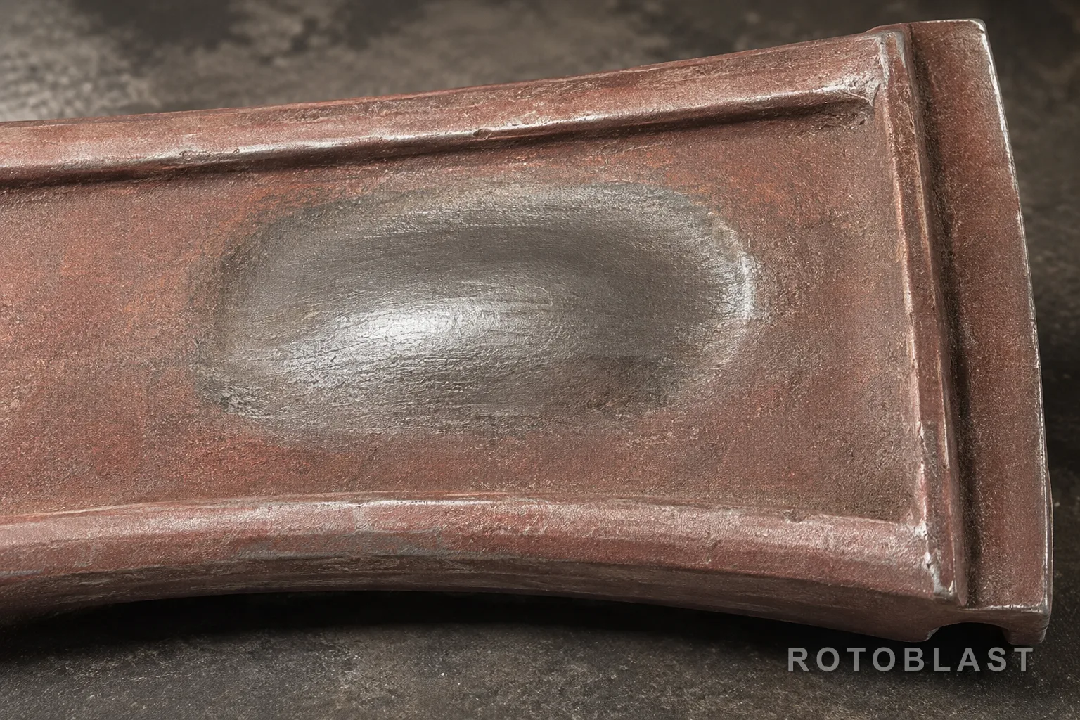 SUBJECT: close-up of a worn liner plate showing a clear hot spot — one dished, polished area with visibly thicker metal around it. FRAMING: tight, the worn patch filling most of the frame, a straight edge included for reference. LIGHTING: hard side light so the dish and the depth difference throw a shadow. ANGLE: shallow, almost across the surface. ENGINEERING STORY: proves wear is concentrated, not spread. PURPOSE: the single most convincing image on the page.